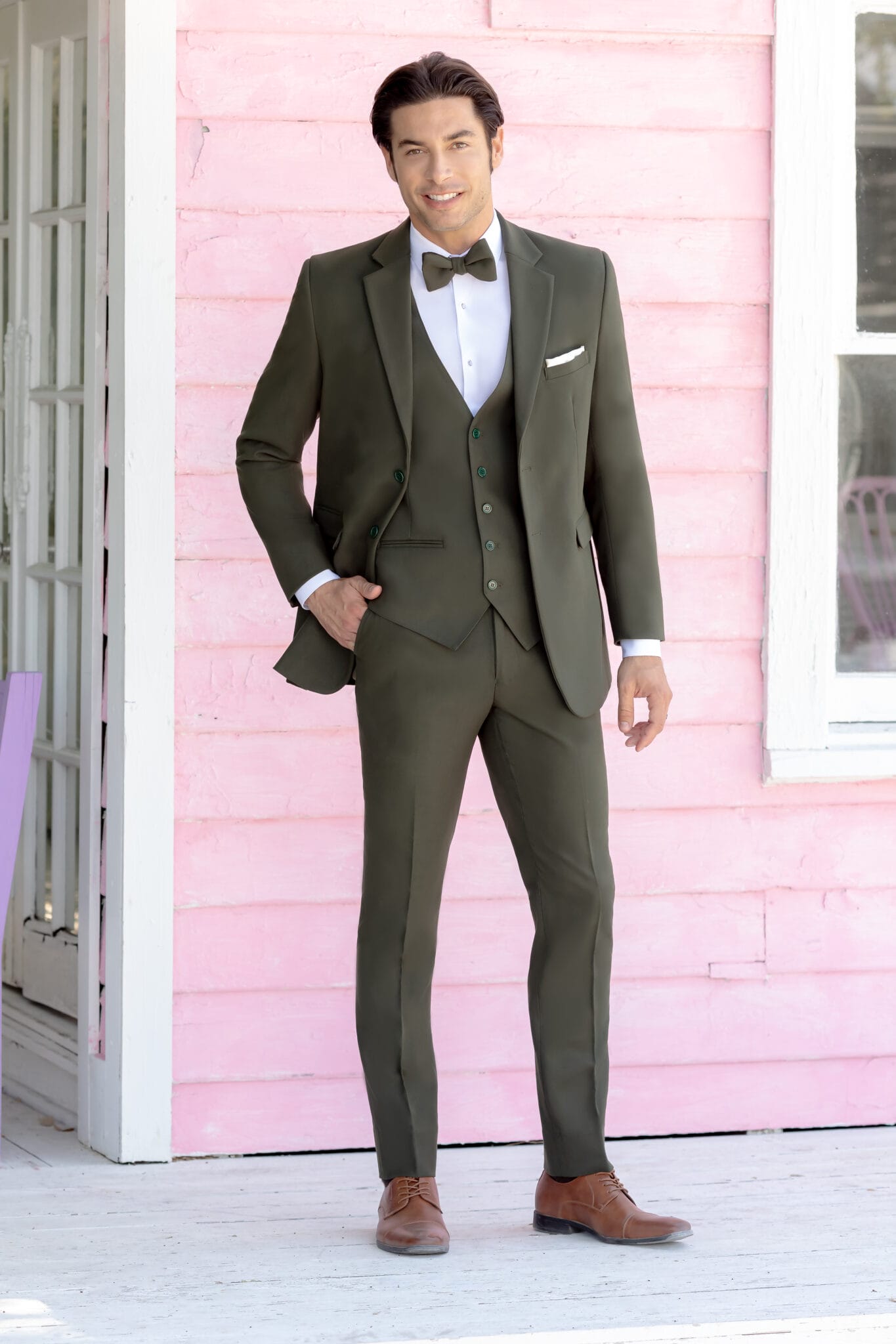 A man in an Indigo Metro Slim Fit Suit with a bow tie stands in front of a pink wooden building, smiling at the camera with one hand in his pocket.