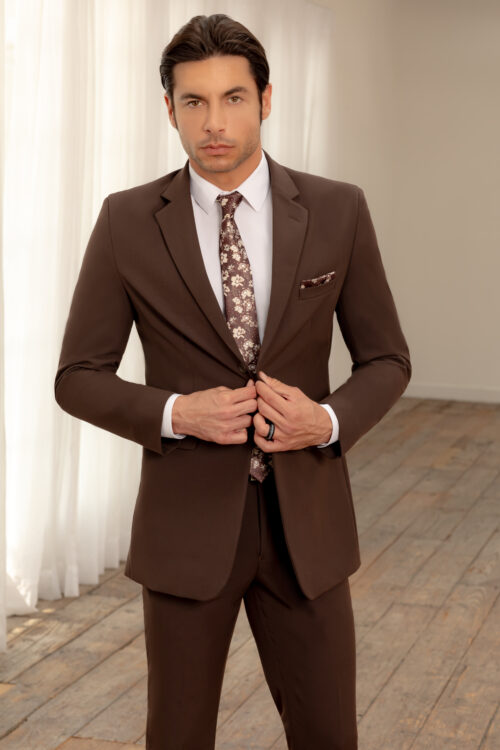 A man stands indoors wearing a Light Grey Metro Slim Fit Suit with a white shirt and a floral tie. He is buttoning the jacket while looking at the camera. The background features white curtains and a wooden floor.