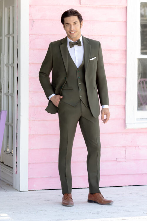 A man in an Indigo Metro Slim Fit Suit with a bow tie stands in front of a pink wooden building, smiling at the camera with one hand in his pocket.