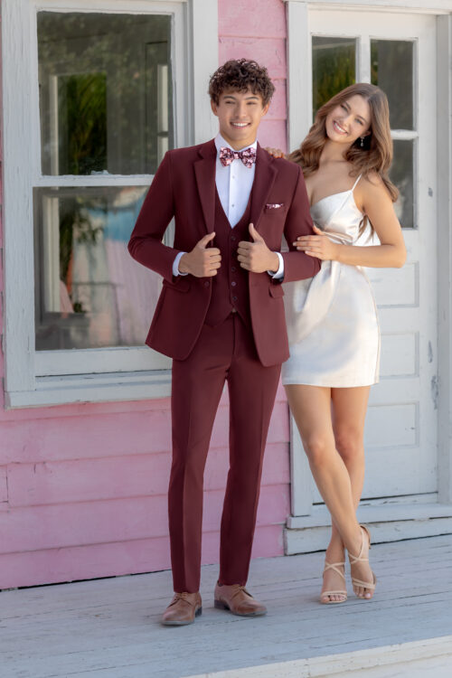 A young man in a Sage Metro Slim Fit Suit and bow tie stands next to a young woman in a white dress. They pose in front of a pink building with white trim and a door.
