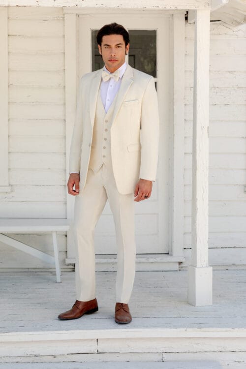 A man in a Burgundy Metro Slim Fit Suit stands in front of a white wooden building and door, with a bench to his left. He is wearing brown shoes and a bow tie.