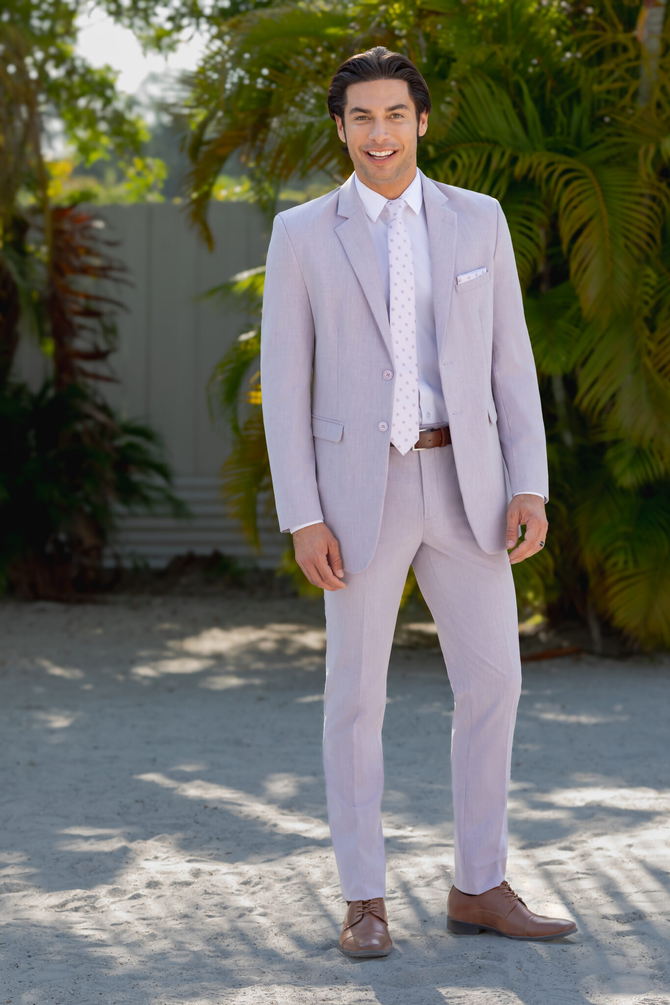 A man stands outdoors in a Charcoal Metro Slim Fit Suit, white shirt, and patterned tie, smiling in front of greenery and a wooden fence.