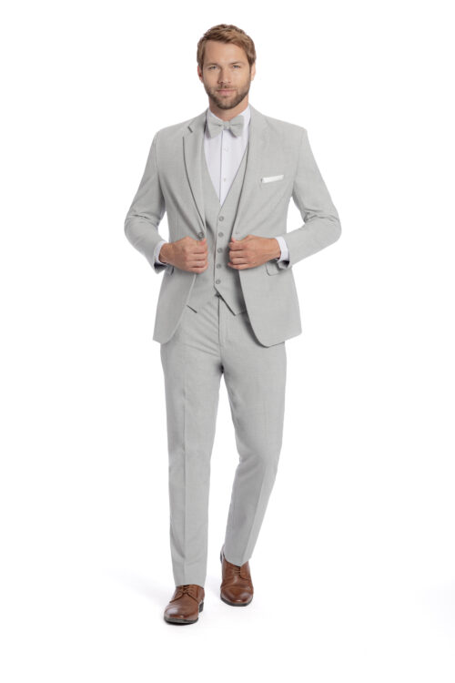 A man in a Green Metro Slim Fit Suit, paired with a bow tie, white dress shirt, and brown shoes, stands with his hands on his jacket.
