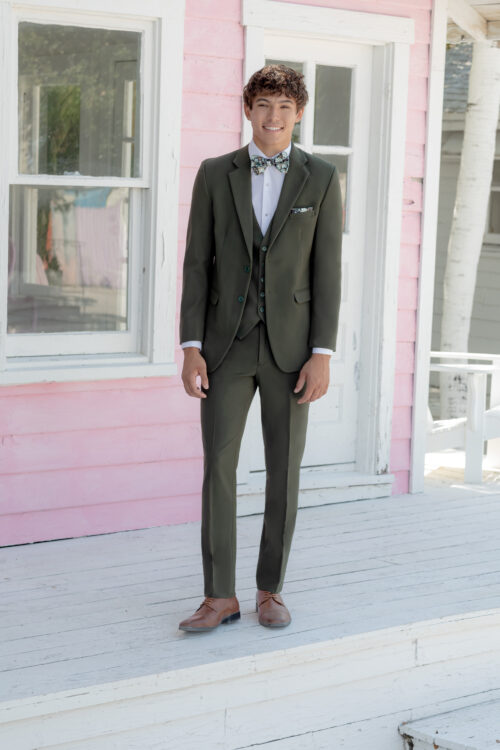 A person in an Indigo Metro Slim Fit Suit, bow tie, and brown shoes is standing on a wooden porch in front of a pink house.