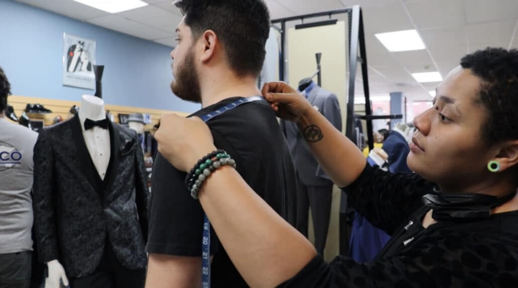 A man is assisting a woman in a clothing store with suit rental options.