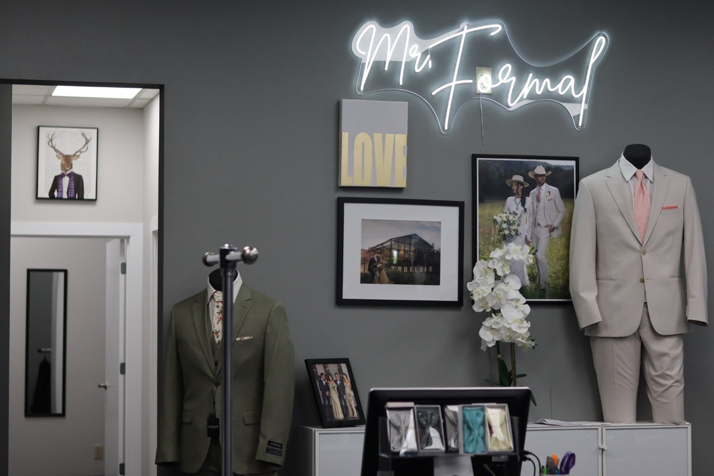 A display of men's formal wear in a store, including a light gray suit and a green jacket on mannequins, with a neon sign that reads "Mr. Formal," and framed pictures on the wall.